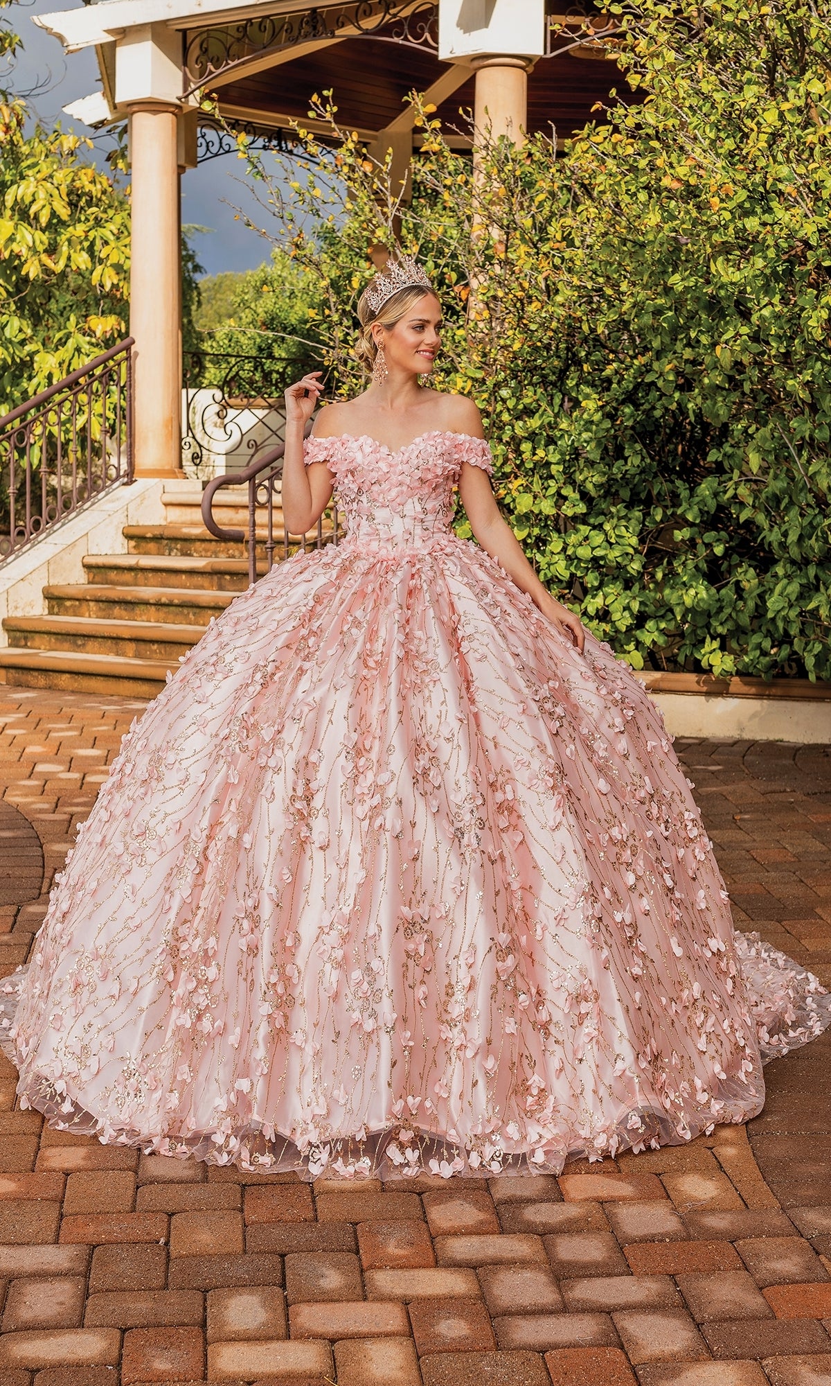 A woman stands outdoors in the Quinceanera Dress 1734 By Dancing Queen, an off-the-shoulder pink ball gown with a sweetheart neckline and floral embellishments, wearing a tiara on a brick path near greenery and a gazebo.